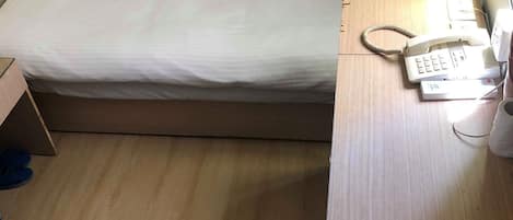 Una cama individual con sábana blanca, una mesita de noche de madera con teléfono y una caja en el suelo.