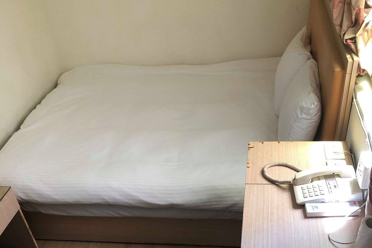 Una cama individual con colchón blanco, una mesita de noche de madera con teléfono y un bulto en el suelo.