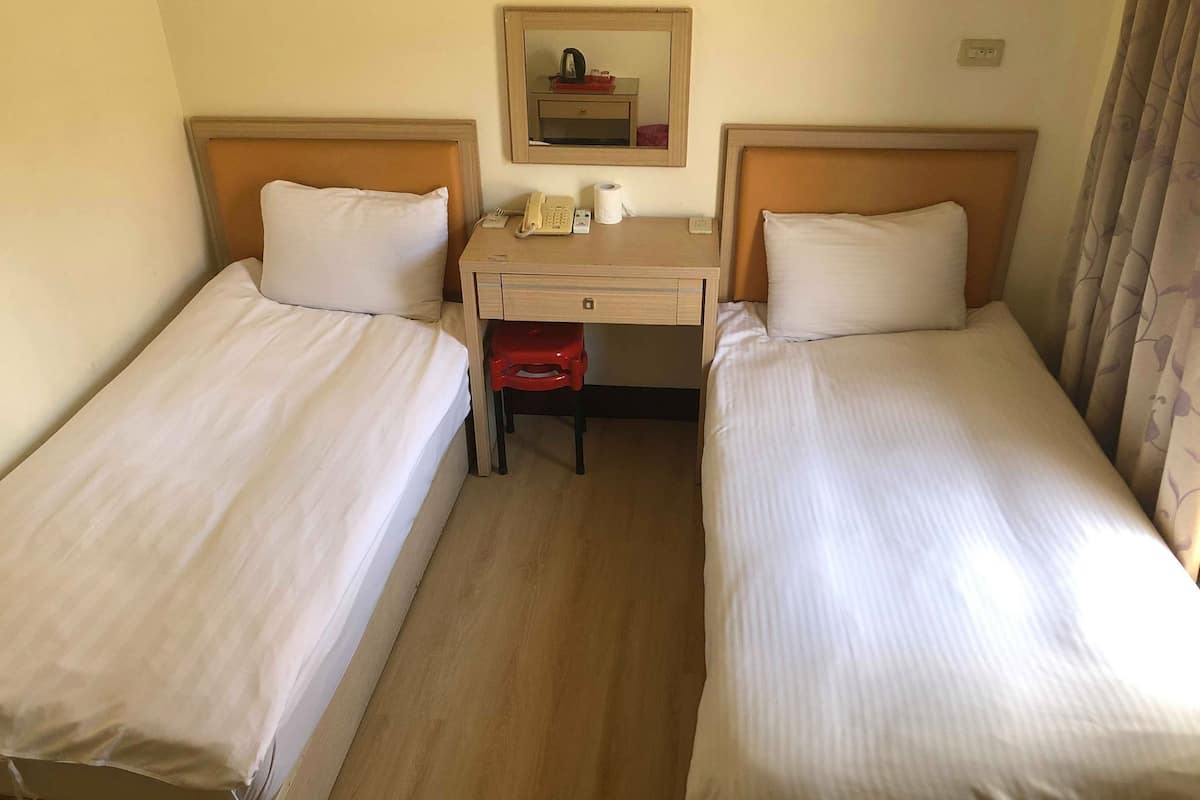 Una habitación de hotel con dos camas individuales, un escritorio con teléfono y una taza, y un espejo en la pared.