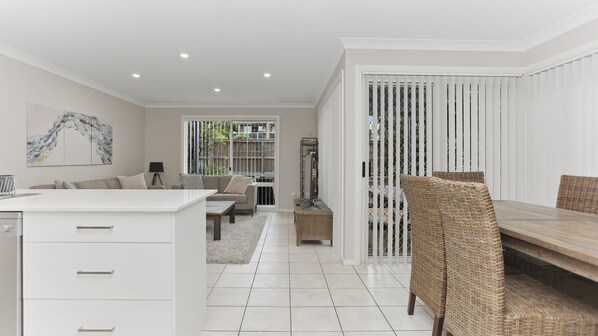 A modern kitchen with white cabinetry, a dining area with wicker chairs, a sofa set, and a view through a glass door to an outdoor space.