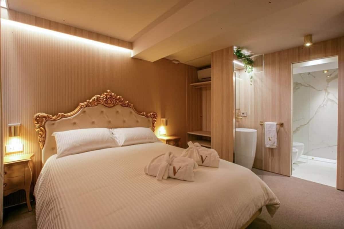 A hotel room with a large bed, bedside tables, a mirror, a wardrobe, and a bathroom with a shower and toilet.