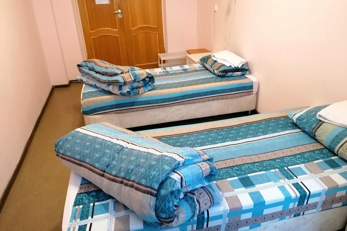 A room with two beds, each with patterned bedding. There is a wooden door and a red lamp on the wall.