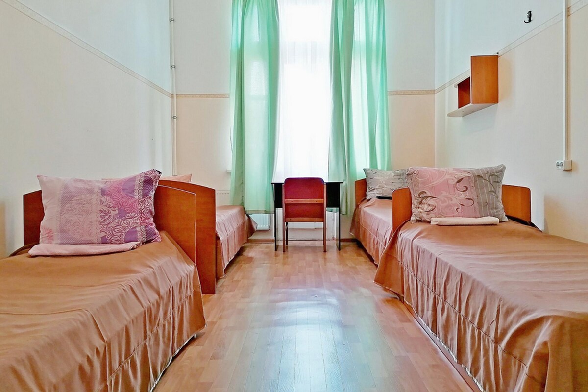 A room with two beds, a chair, and a window with green curtains.