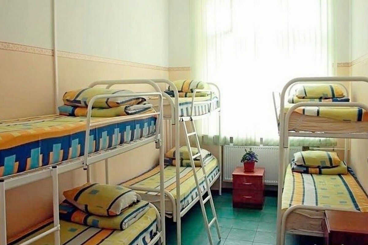 A dormitory room with bunk beds, a small red cabinet, and a window with curtains.