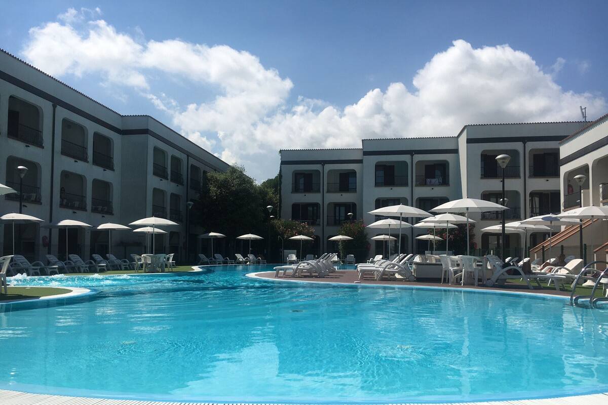 A swimming pool with lounge chairs and umbrellas, surrounded by multi-story buildings with balconies.