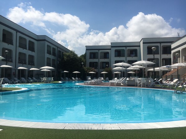 Una piscina con sillas de descanso y sombrillas, rodeada de edificios de varios pisos con balcones.