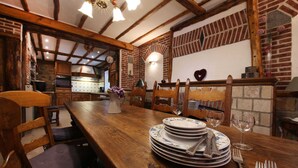 A rustic dining area with a wooden table set for four, surrounded by chairs. A stack of plates and cutlery is visible, and a vase with purple flowers adds a touch of color. The room features exposed wooden beams and brick walls, creating a cozy atmosphere. - Family House with Large Garden & Games Room near Bambois Lake (Mettet)