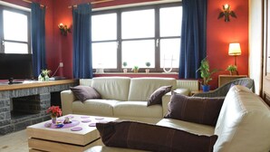 Un salon avec un canapé beige, une table basse en bois et une cheminée en pierre. Il y a deux fenêtres avec des rideaux bleus, un téléviseur et une lampe sur une table d’appoint.