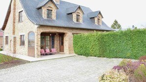 Une grande maison en pierre, dotée d’un toit à forte pente, d’une cheminée en briques et d’une haie bien entretenue.