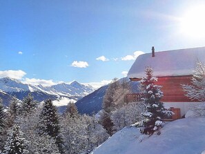 A snow-covered mountain landscape with a red building featuring a chimney, surrounded by snow-laden trees and mountains in the background.
