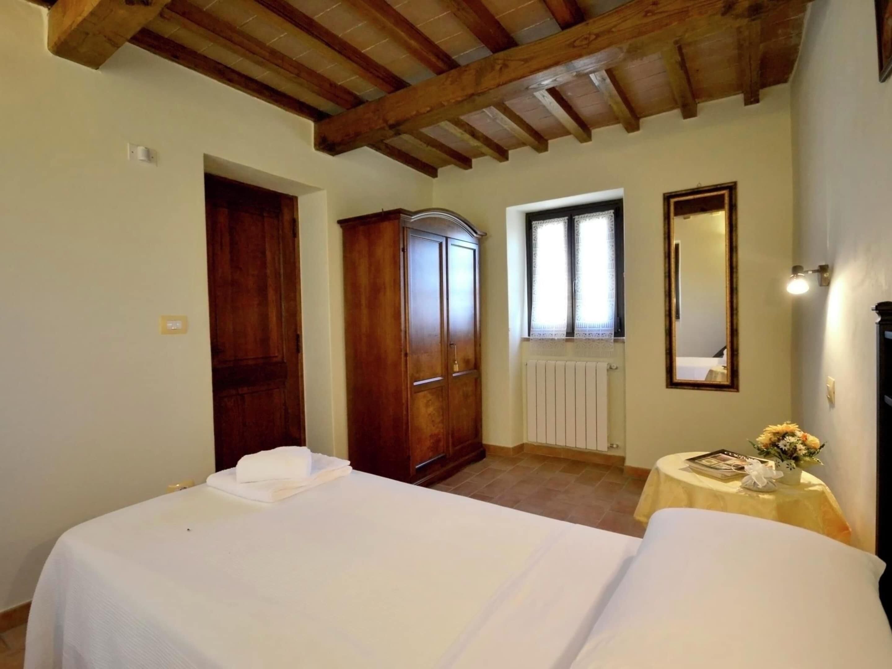 A bedroom with two beds, a wooden wardrobe, a radiator, a window with curtains, a round table with a vase, and a mirror on the wall.