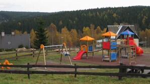 A playground with swings, slides, and climbing structures, surrounded by a wooden fence and a forested hillside in the background. - Lavish Holiday Home in Abertamy near Ski Area (Abertamy)