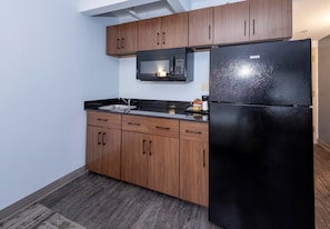 A compact kitchen with wooden cabinets, a black refrigerator, a microwave, and a sink. - HomeTowne Studios by Red Roof Wilmington - New Castle (New Castle)