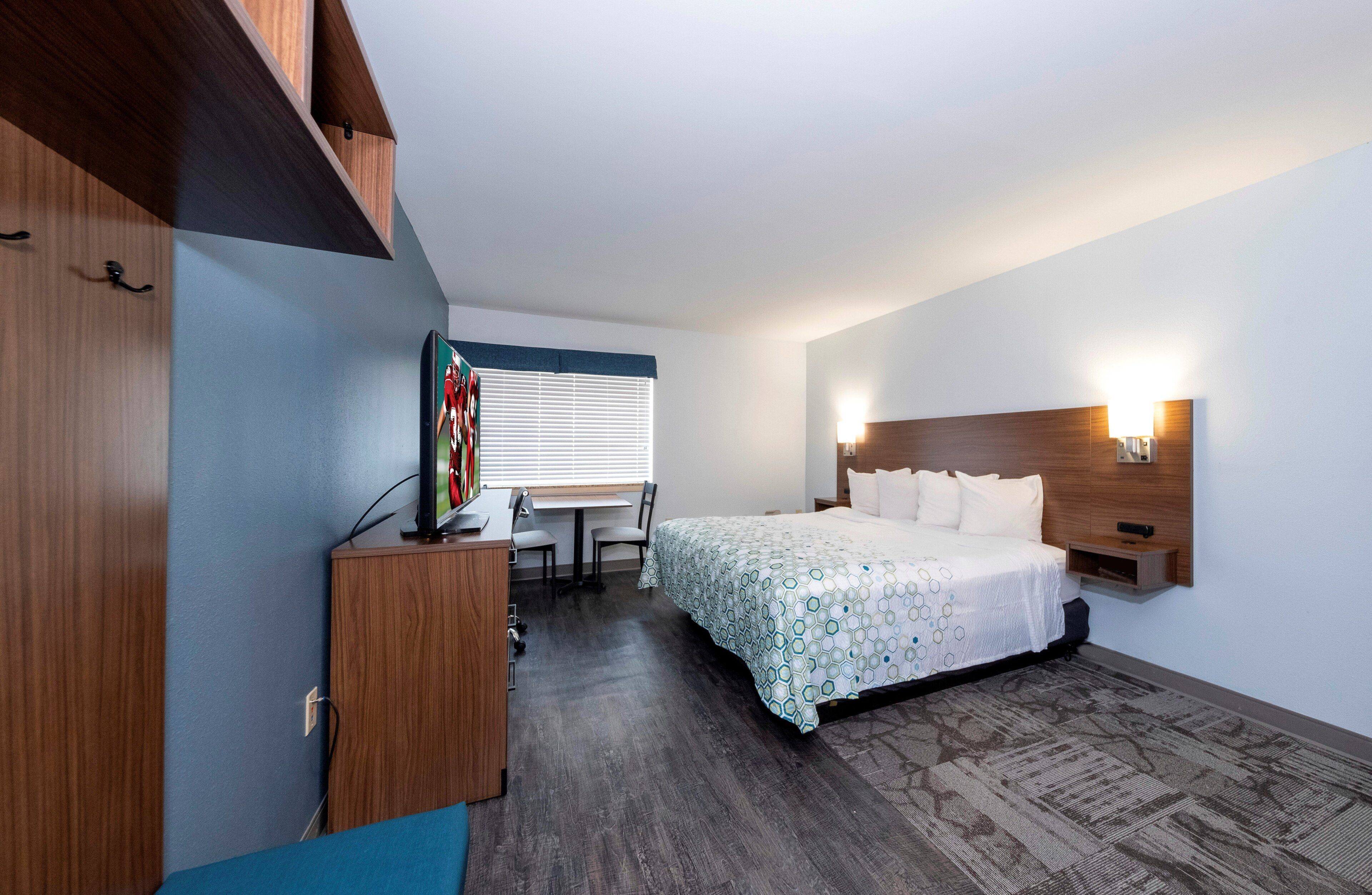 Habitación de hotel con cama, escritorio, silla, televisor y ventana con persianas.