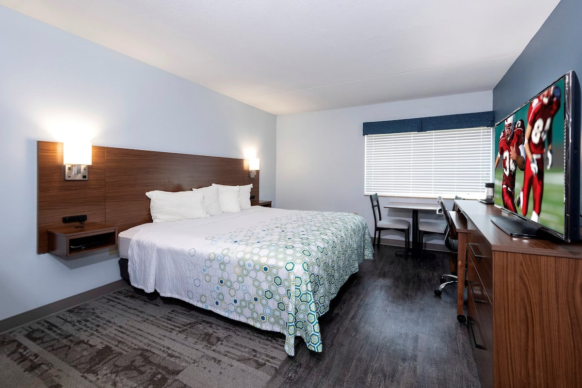 A hotel room with a bed, bedside tables, a desk, a chair, a television, and a window with blinds.