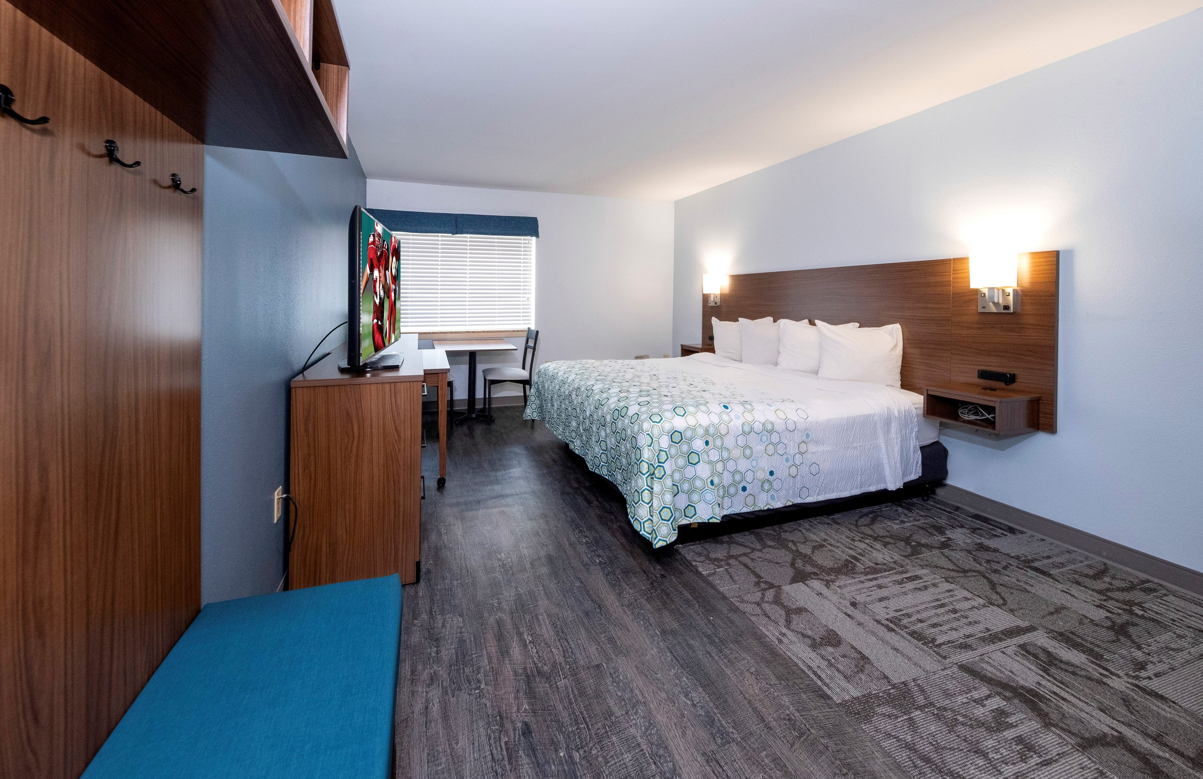 A hotel room with a bed, a desk, a chair, a TV, and a wall-mounted light.