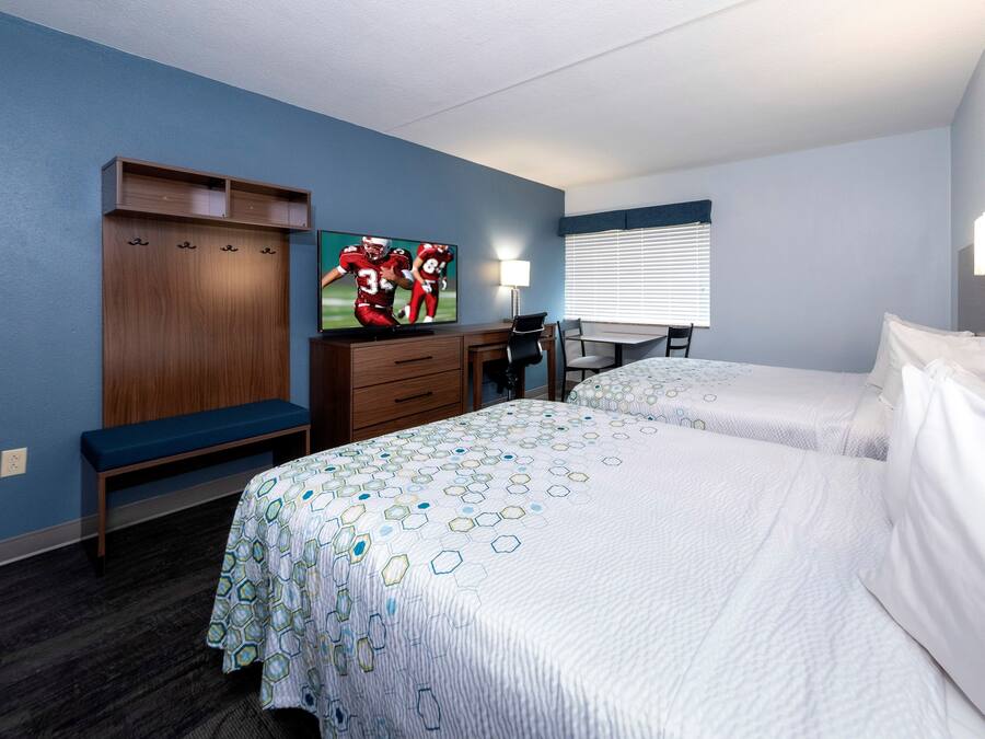 Habitación de hotel con dos camas, un escritorio con silla, televisor y ventana con persianas.