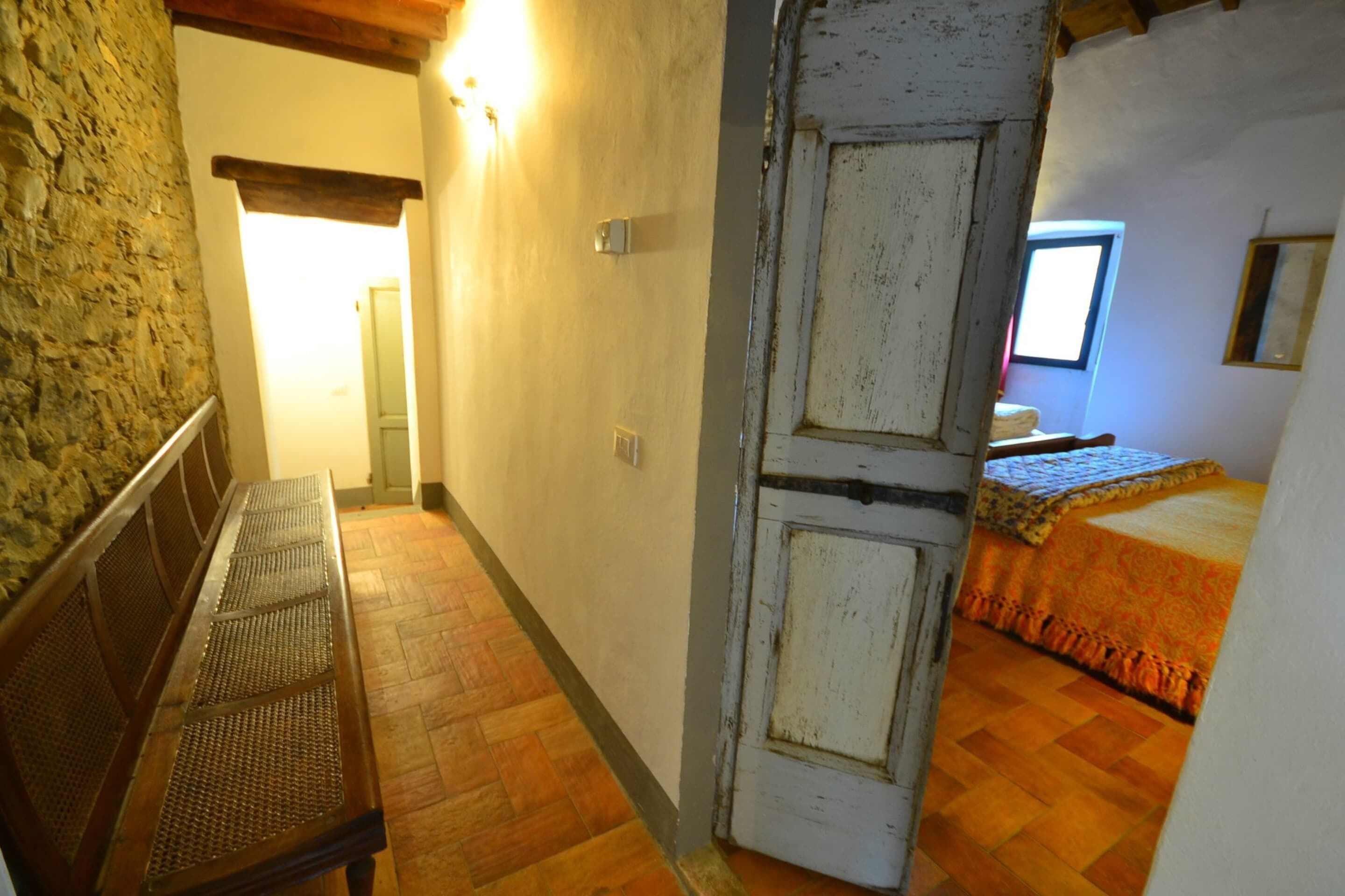 Un couloir étroit avec un banc en bois, une porte blanche et un lit recouvert d’une couverture orange.