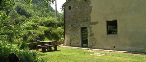 A rustic stone building with a grassy yard, a wooden bench, and a stone wall.