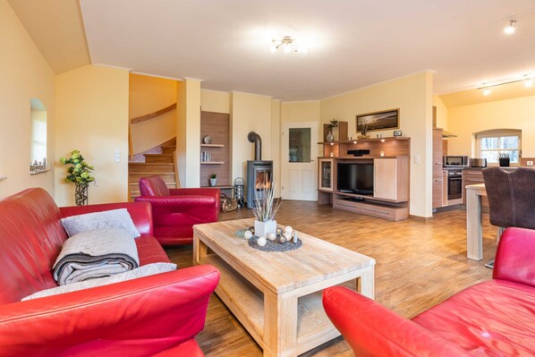 A spacious living area with a wooden coffee table, red leather sofas, a TV, and a kitchen in the background.
