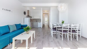 Ein modernes Wohnzimmer mit Sofa, Esstisch und Stühlen. Im Hintergrund ist eine Küche zu sehen.