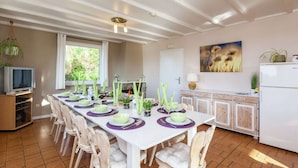 Une salle à manger avec une longue table dressée pour six personnes, entourée de chaises en bois. Un téléviseur est posé sur une étagère à gauche, et une cheminée est visible au fond.