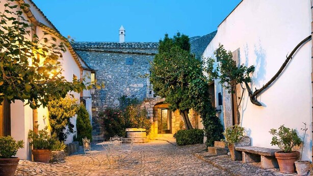 Una calle adoquinada en un pueblo histórico con edificios de piedra, un muro de piedra y plantas en macetas.