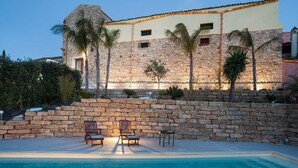 Edificio de piedra con piscina, dos sillones de descanso y palmeras.