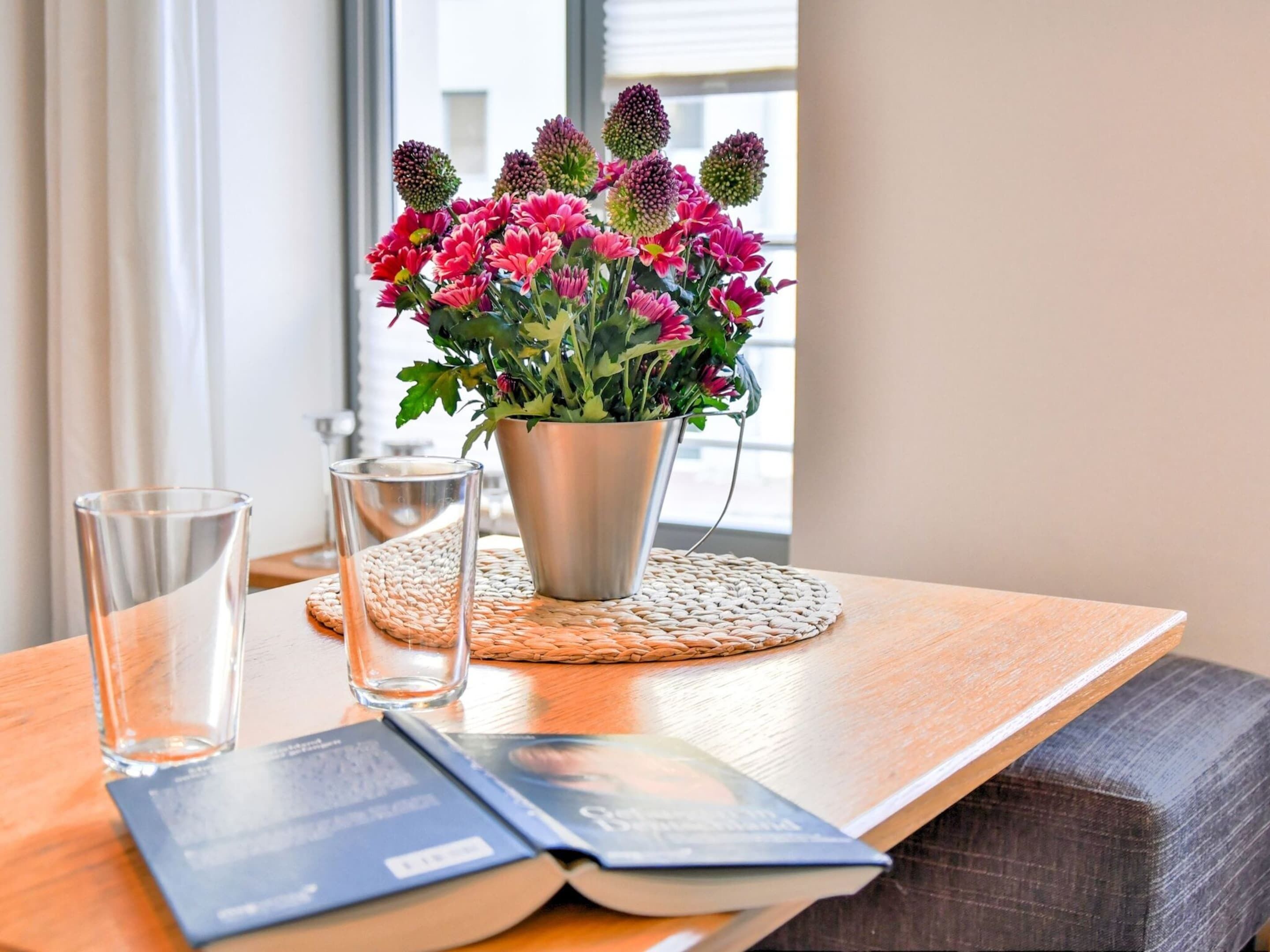 Uma mesa de madeira com um vaso de flores de metal, dois copos e um livro aberto.