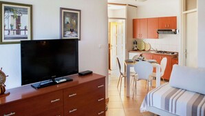Ett kompakt bostadsutrymme med en TV på ett träskåp, en matplats med bord och stolar samt ett litet kök med skåp och en spis.