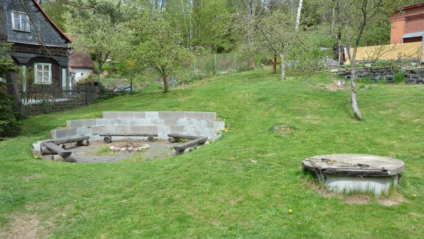 Un jardín trasero con césped, un pozo de fuego de piedra y una estructura de hormigón.