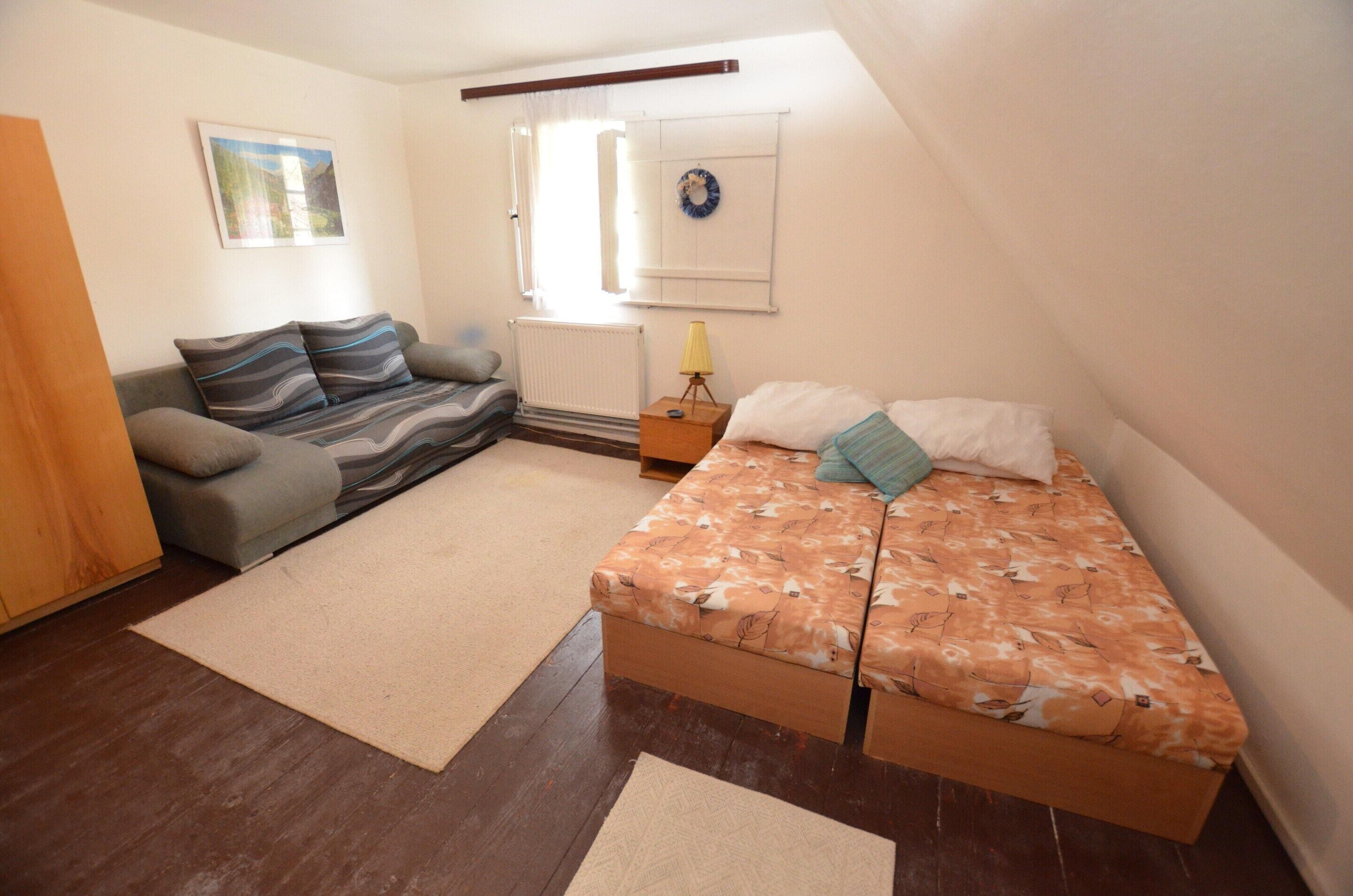 A small, cozy room with a sofa, a bed, a radiator, and a window with curtains.