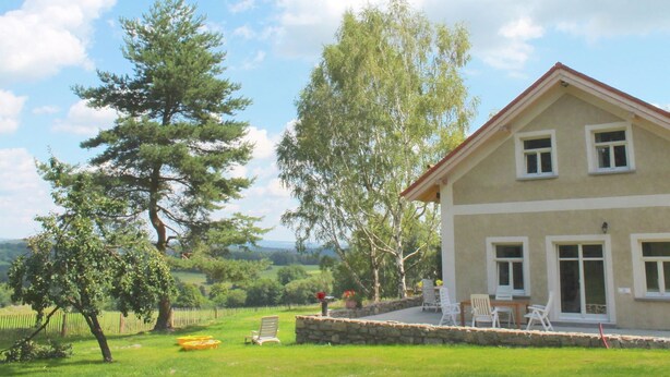 Una casa de dos pisos con base de piedra, un patio con mobiliario exterior y vistas del paisaje circundante.