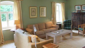 Una sala de estar con un sofá, una mesa de centro y un sillón. Hay dos ventanas con cortinas, una lámpara y cuadros enmarcados en la pared.