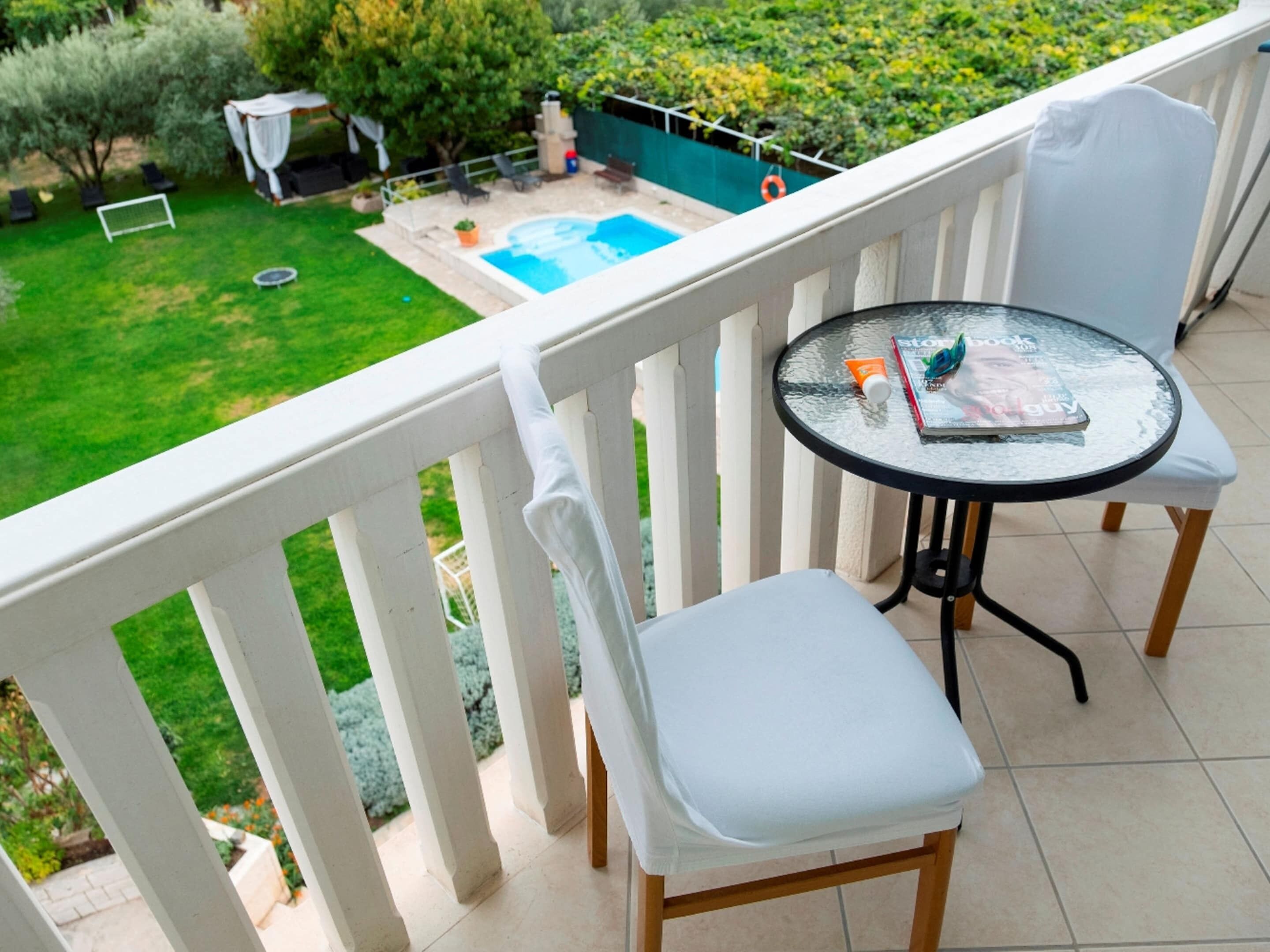 Un balcón con una mesa de vidrio, dos sillas blancas y vistas a una piscina y jardín.