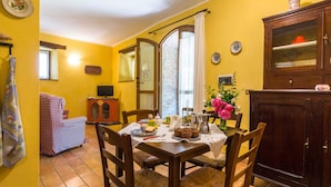 A dining area with a table set for four, surrounded by wooden chairs with woven seats. A cabinet with glass doors is to the right, and a television is on a wooden cabinet to the left. - Belvilla by Oyo Azalea in Gubbio (Gubbio)
