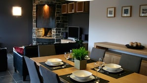 A modern dining area with a stone fireplace, wooden furniture, and a large hanging light fixture. - Robertville Lakeview Escape (Waimes)