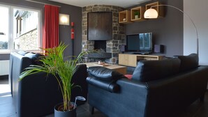 A modern living room with a stone fireplace, leather sofas, a flat-screen TV, and a large window with red curtains. - Robertville Lakeview Escape (Waimes)