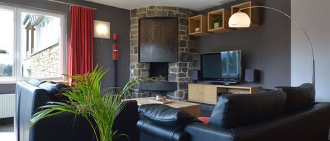 A modern living room with a stone fireplace, leather sofas, a flat-screen TV, and a large window with red curtains.