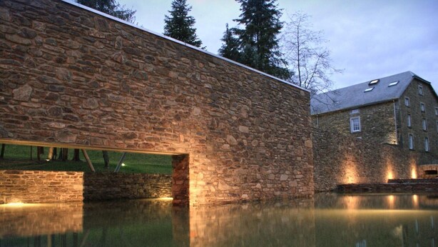 A stone building with a reflective pool in front, a wooden bridge, and a large building in the background. - Grand Retreat in Redu (Libin)