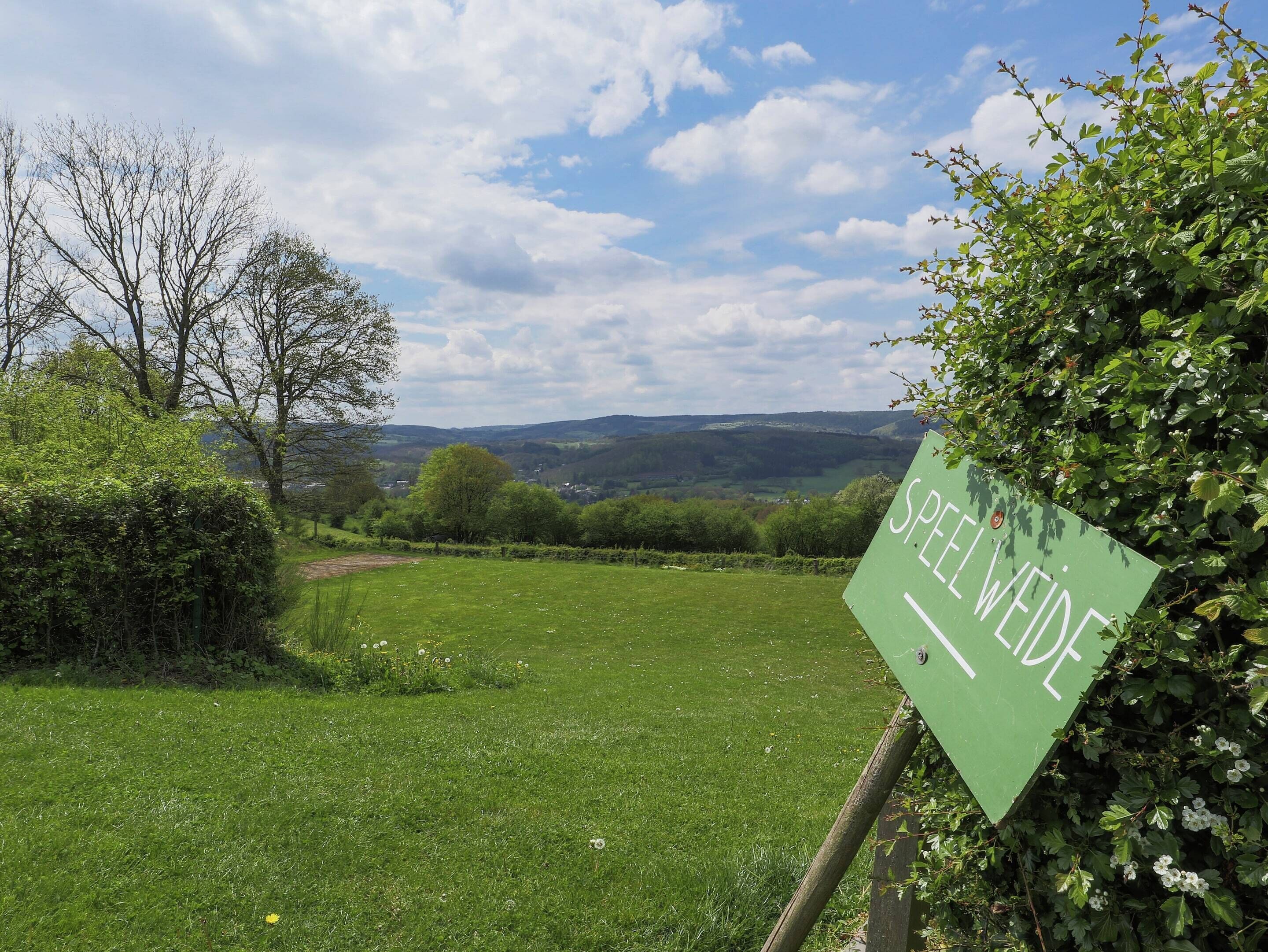 A sign with the text 'SPELWEIDE' in a natural landscape with grass, trees, and a distant view of hills.