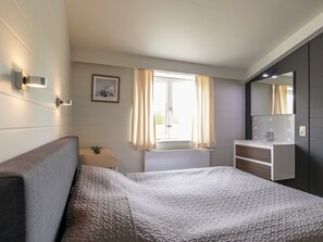 A bedroom with a bed, a bedside table, a window with curtains, a wall-mounted light, and a mirror.