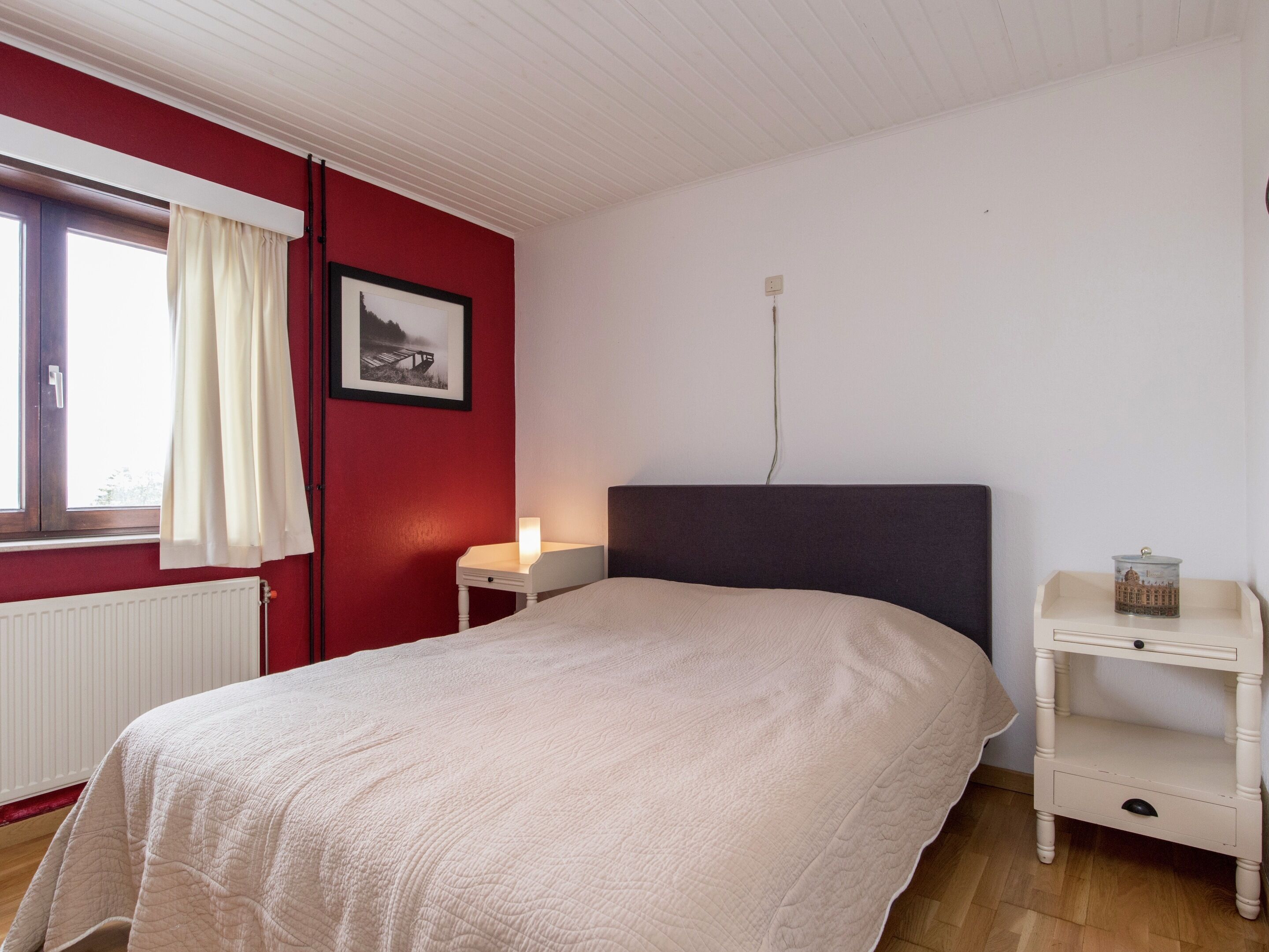 A bedroom with a bed, a nightstand, a lamp, a picture on the wall, and a window with curtains.