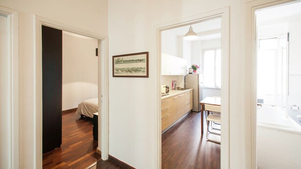 Un corridoio con pavimento in legno, un letto, un angolo cottura con mobili e frigorifero, e un'area pranzo con tavolo e sedie.