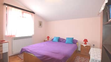 A bedroom with a large bed, a radiator, a window with curtains, a bedside table with a lamp, and a small table with a tablecloth.