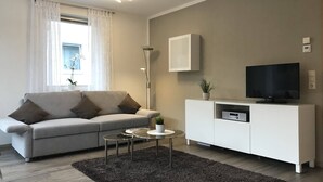 Un salón moderno con un sofá gris, un televisor de pantalla plana sobre un mueble blanco, una lámpara de pie y una pequeña mesita con una planta.
