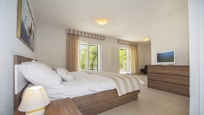 Une chambre à coucher comprenant un lit, une lampe de chevet, une commode, un téléviseur et des fenêtres avec des rideaux.