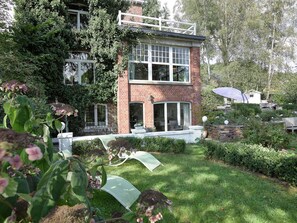 A two-story brick house with a large glass window and door, surrounded by lush greenery and a well-maintained garden. - Stavelot Sauna & Garden Stay (Stavelot)
