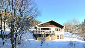 Eine zweistöckige Chalet mit Natursteinfassade und Holzbalkon, umgeben von schneebedeckten Bäumen und blauem Himmel.