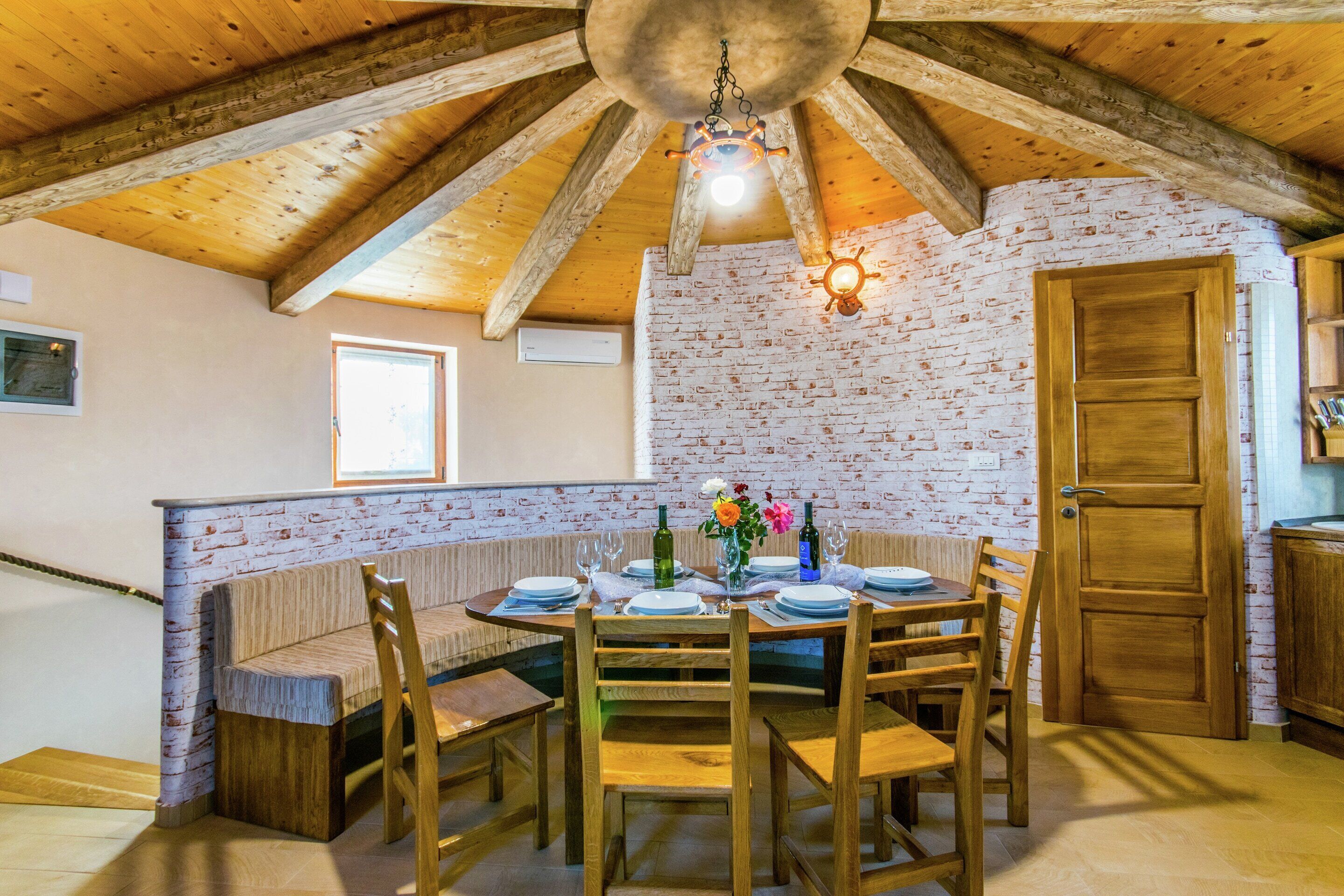 A dining area with a brick wall, wooden ceiling beams, and a round table set for six.